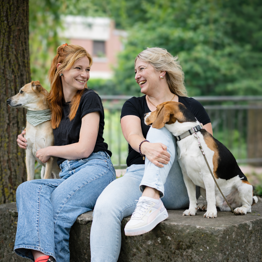 Merle und Melissa sitzen lachend mit ihren Therapiebegleithunden Lio und Neo im Grünen.